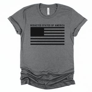 K&ouml;nnte beinhalten: Ein graues T-Shirt mit einem schwarzen Flaggen-Design. Die Flagge hat horizontale Streifen und ein durchgehendes Rechteck in der oberen linken Ecke. Der Text "REDACTED STATES OF AMERICA" ist &uuml;ber der Flagge aufgedruckt. Das T-Shirt hat einen Rundhalsausschnitt und kurze &Auml;rmel.