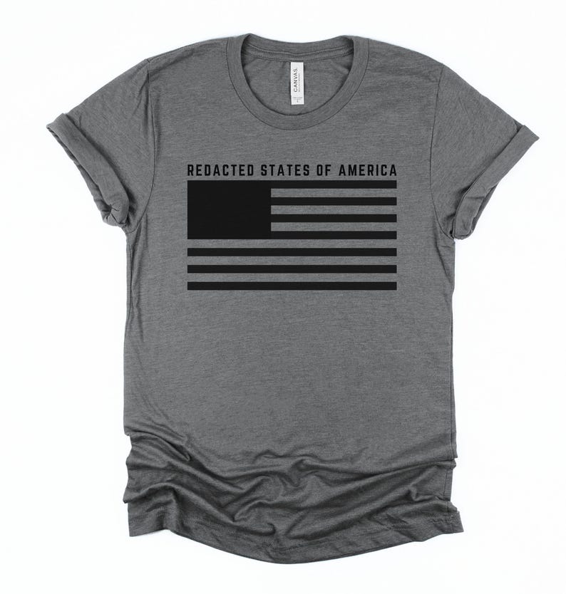 K&ouml;nnte beinhalten: Ein graues T-Shirt mit einem schwarzen Flaggen-Design. Die Flagge hat horizontale Streifen und ein durchgehendes Rechteck in der oberen linken Ecke. Der Text "REDACTED STATES OF AMERICA" ist &uuml;ber der Flagge aufgedruckt. Das T-Shirt hat einen Rundhalsausschnitt und kurze &Auml;rmel.