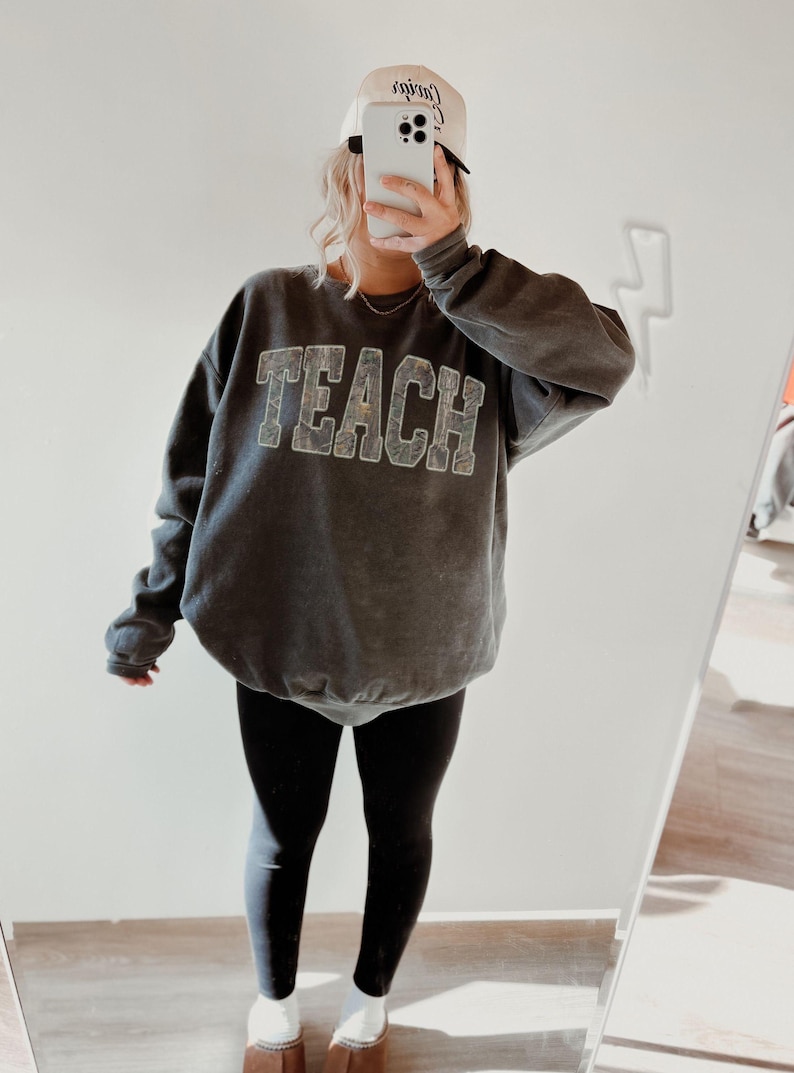 May include: A gray sweatshirt with the word "TEACH" in large, textured letters. The person is wearing black leggings, a white baseball cap, and brown clogs. The cap has text on it.