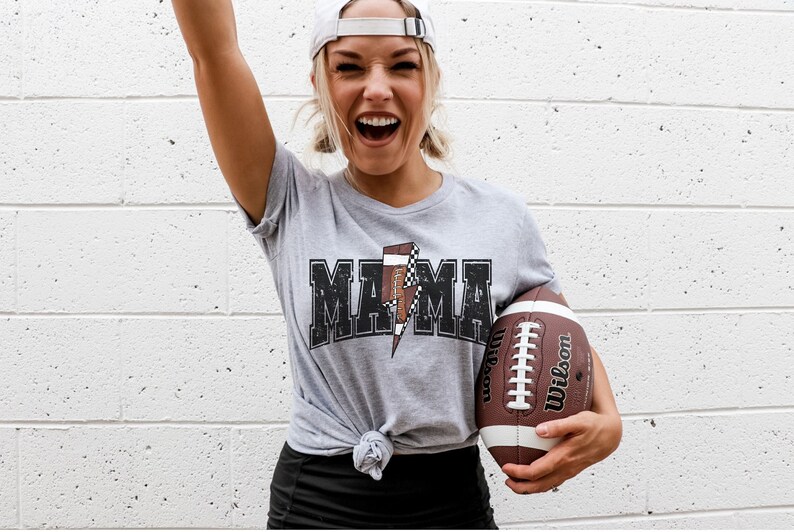 May include: A woman wearing a grey t-shirt with a black "MAMA" graphic and a lightning bolt design. The lightning bolt is made up of a football and a checkered pattern. She is holding a brown football.