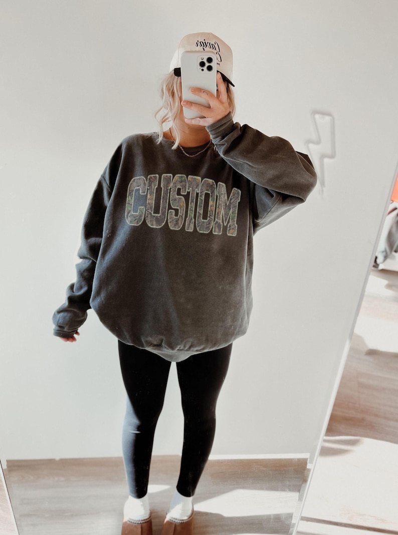 K&ouml;nnte beinhalten: Ein graues Sweatshirt mit dem Wort "CUSTOM" in einer verwaschenen, strukturierten Schriftart. Die Person tr&auml;gt schwarze Leggings, eine wei&szlig;e Baseballkappe und braune Hausschuhe. Das Bild wird vor einem Spiegel aufgenommen.