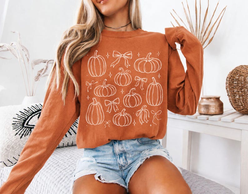 May include: A long-sleeve crewneck sweatshirt in a burnt orange color featuring a white line-art graphic print. The design consists of various pumpkin shapes interspersed with decorative bows and small four-pointed star accents across the front.