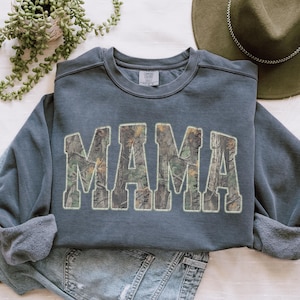 May include: A slate blue sweatshirt with the word "MAMA" in large, block letters filled with a camouflage pattern. The sweatshirt is folded, with the sleeves and bottom hem visible. A green hat and jeans are in the background.