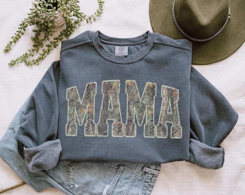 May include: A slate blue sweatshirt with the word "MAMA" in large, block letters filled with a camouflage pattern. The sweatshirt is folded, with the sleeves and bottom hem visible. A green hat and jeans are in the background.