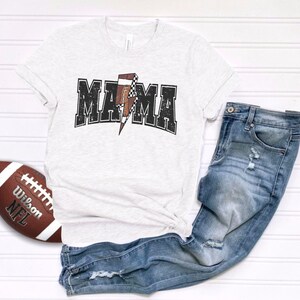 May include: A white t-shirt with a distressed black graphic that says "MAMA" with a football-shaped lightning bolt in the middle. The shirt is paired with blue jeans.