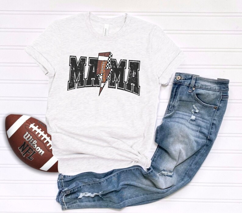 May include: A white t-shirt with a distressed black graphic that says "MAMA" with a football-shaped lightning bolt in the middle. The shirt is paired with blue jeans.
