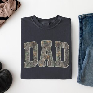 May include: A charcoal gray crewneck sweatshirt featuring the word DAD in large, block-style letters. The letters are filled with a realistic forest camouflage pattern in shades of green, brown, and tan, with a light-colored outline around each letter.