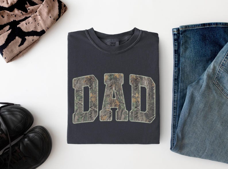 May include: A charcoal gray crewneck sweatshirt featuring the word DAD in large, block-style letters. The letters are filled with a realistic forest camouflage pattern in shades of green, brown, and tan, with a light-colored outline around each letter.