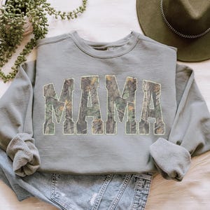 May include: A sage green sweatshirt with the word "MAMA" in large, block letters filled with a camouflage pattern. The sweatshirt is folded on top of a pair of blue jeans. A green hat and a potted plant are in the background.