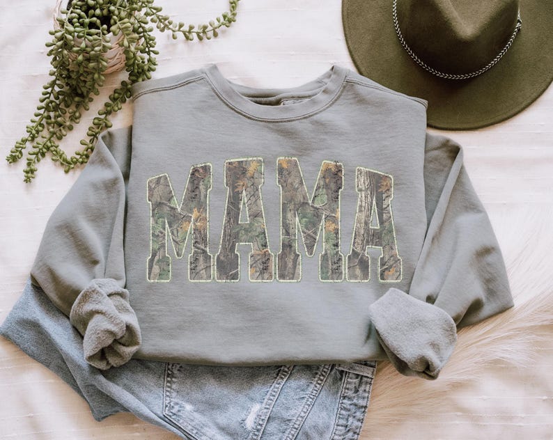 May include: A sage green sweatshirt with the word "MAMA" in large, block letters filled with a camouflage pattern. The sweatshirt is folded on top of a pair of blue jeans. A green hat and a potted plant are in the background.