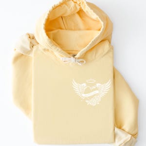 May include: A folded pale yellow hoodie with a drawstring hood and a white graphic on the front. The graphic features a heart with wings, a halo, and roses. The hoodie is made of a soft material.