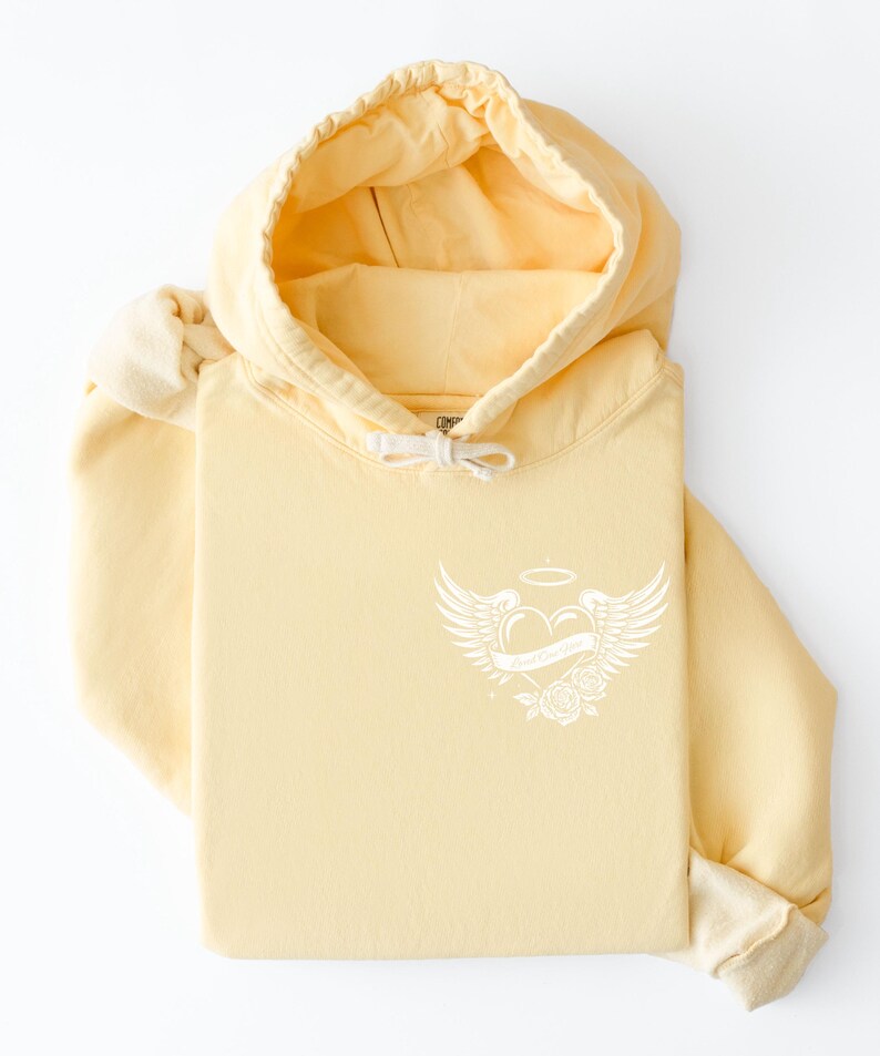 May include: A folded pale yellow hoodie with a drawstring hood and a white graphic on the front. The graphic features a heart with wings, a halo, and roses. The hoodie is made of a soft material.