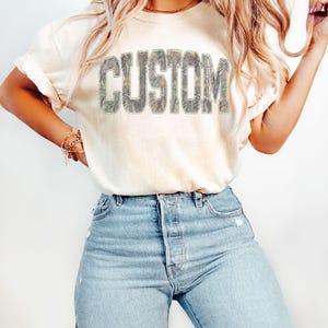 K&ouml;nnte beinhalten: Cremefarbenes T-Shirt mit dem Wort "CUSTOM" in einem Camouflage-Design auf der Brust. Das Shirt wird mit hellblauen Jeans kombiniert. Das T-Shirt hat hochgekrempelte &Auml;rmel und eine lockere Passform.