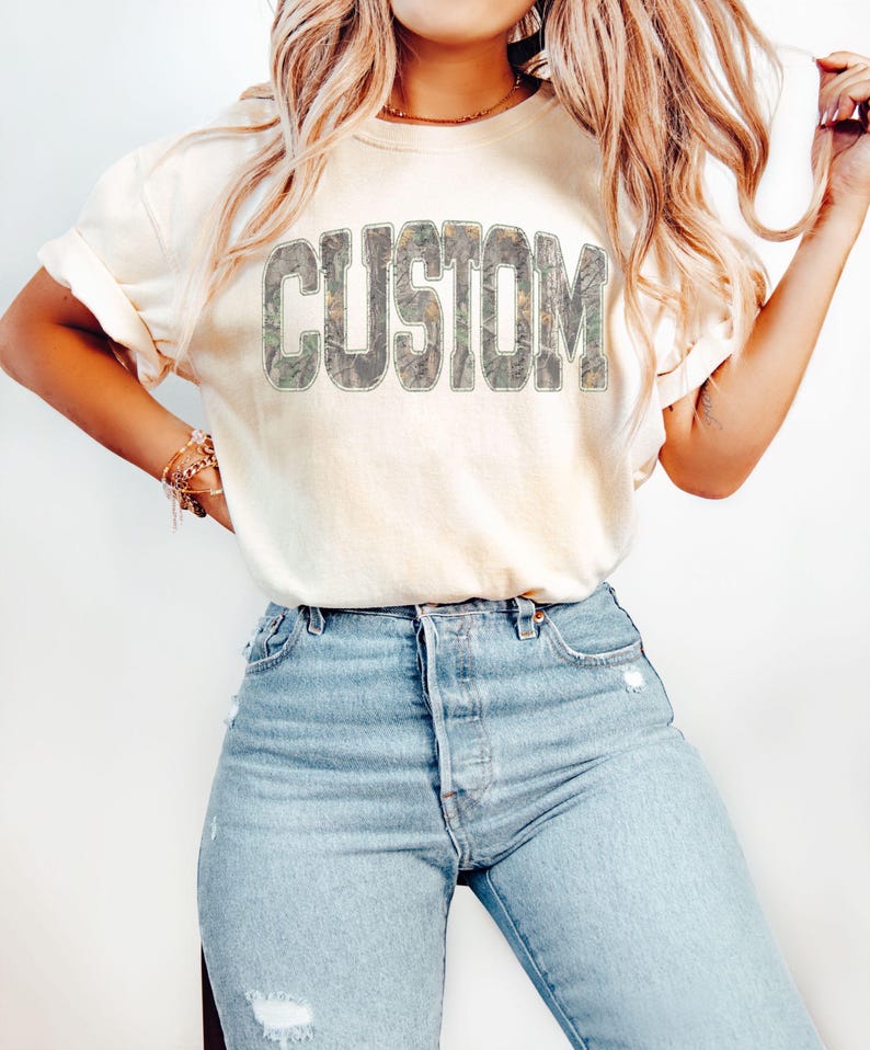 K&ouml;nnte beinhalten: Cremefarbenes T-Shirt mit dem Wort "CUSTOM" in einem Camouflage-Design auf der Brust. Das Shirt wird mit hellblauen Jeans kombiniert. Das T-Shirt hat hochgekrempelte &Auml;rmel und eine lockere Passform.