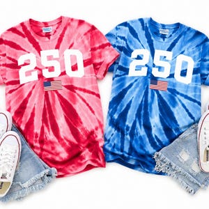 May include: Two tie-dye t-shirts, one red and one blue, each with the number "250" in white and an American flag patch. White trainers and denim shorts are paired with each shirt. American flags are placed on the sides.