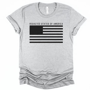 K&ouml;nnte beinhalten: Hellgraues T-Shirt mit einer schwarzen Grafik einer zensierten amerikanischen Flagge. Der Text "REDACTED STATES OF AMERICA" befindet sich &uuml;ber dem Flaggen-Design. Das Shirt hat kurze &Auml;rmel und einen Rundhalsausschnitt.