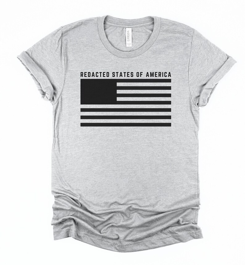 K&ouml;nnte beinhalten: Hellgraues T-Shirt mit einer schwarzen Grafik einer zensierten amerikanischen Flagge. Der Text "REDACTED STATES OF AMERICA" befindet sich &uuml;ber dem Flaggen-Design. Das Shirt hat kurze &Auml;rmel und einen Rundhalsausschnitt.