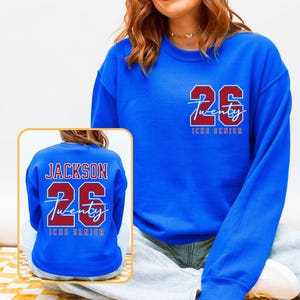 May include: A royal blue sweatshirt with the number "26" and the word "Twenty" in a red and white design. The text "ICHS Senior" is below the number. The back of the sweatshirt also features the name "Jackson" and the number "26".