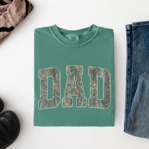 May include: A sage green crewneck t-shirt featuring the word DAD printed in a block font with a camouflage pattern. The shirt is displayed alongside a pair of blue denim jeans, a pair of black sneakers, and a tan and black patterned beanie.