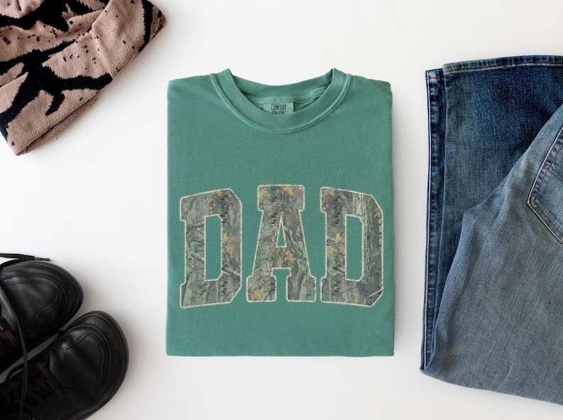 May include: A sage green crewneck t-shirt featuring the word DAD printed in a block font with a camouflage pattern. The shirt is displayed alongside a pair of blue denim jeans, a pair of black sneakers, and a tan and black patterned beanie.
