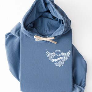 May include: A blue hoodie with a drawstring and a white graphic on the front. The graphic features a heart with wings, a halo, and roses, with the text "Loved One Here". The hoodie is folded.