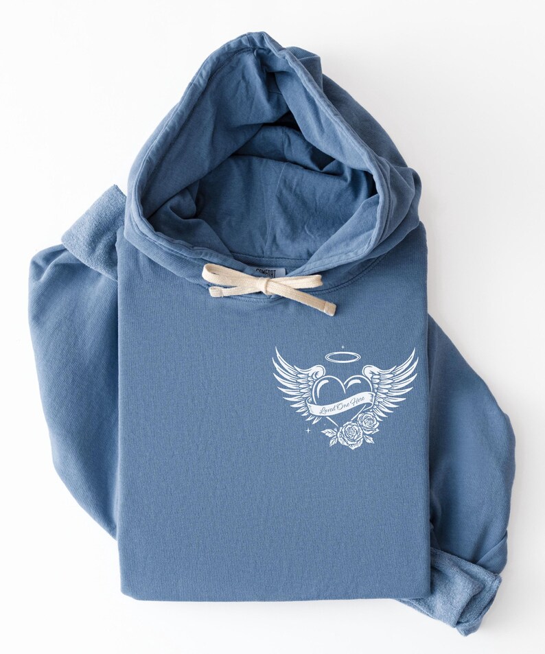 May include: A blue hoodie with a drawstring and a white graphic on the front. The graphic features a heart with wings, a halo, and roses, with the text "Loved One Here". The hoodie is folded.