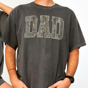 May include: A charcoal gray short-sleeve t-shirt featuring the word DAD printed in large block letters across the chest. The letters are filled with a realistic forest camouflage pattern in shades of brown, green, and tan, outlined in white.