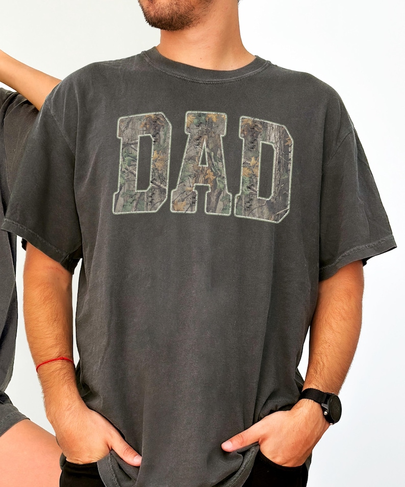 May include: A charcoal gray short-sleeve t-shirt featuring the word DAD printed in large block letters across the chest. The letters are filled with a realistic forest camouflage pattern in shades of brown, green, and tan, outlined in white.