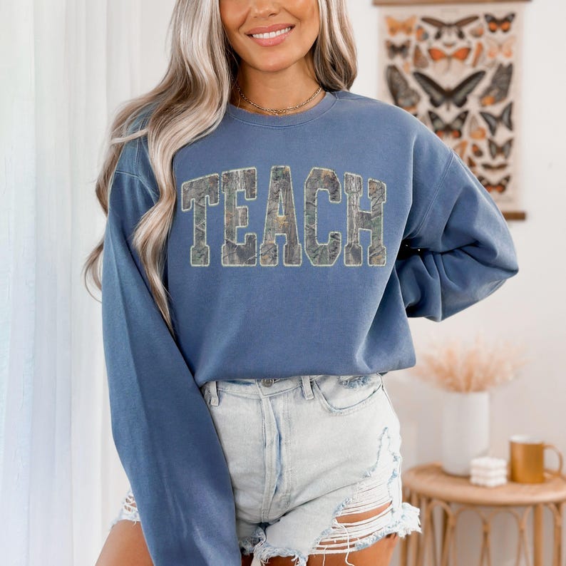 K&ouml;nnte beinhalten: Ein blaues Langarm-Sweatshirt mit dem Wort "TEACH" in einem Camouflage-Muster auf der Brust. Das Sweatshirt wird mit Jeansshorts kombiniert. Der Hintergrund umfasst einen dekorativen Wandbehang und einen kleinen Tisch.