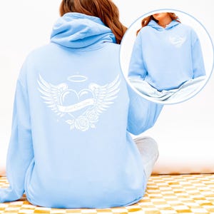 May include: A light blue hoodie with a white graphic on the back featuring a heart with wings, a banner that reads "Loved One Here", and roses. The hoodie has a drawstring hood and a kangaroo pocket.