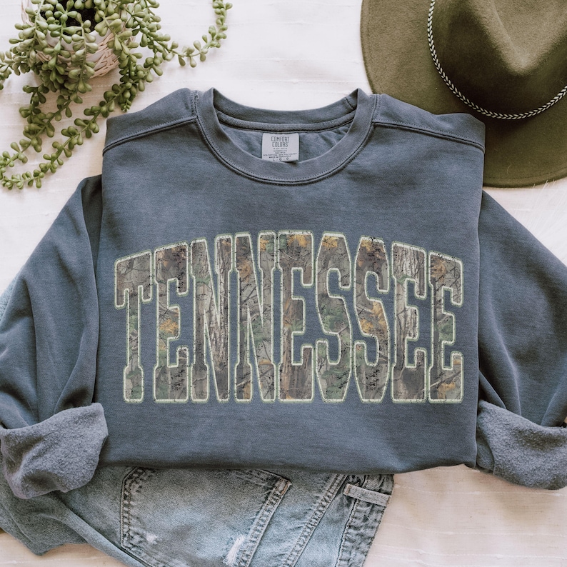 K&ouml;nnte beinhalten: Ein schieferblaues Sweatshirt mit dem Wort "TENNESSEE" in gro&szlig;en, bogenf&ouml;rmigen Buchstaben. Die Buchstaben sind mit einem Tarnmuster aus Gr&uuml;n-, Braun- und Beiget&ouml;nen gef&uuml;llt. Das Sweatshirt hat einen Rundhalsausschnitt und lange &Auml;rmel.