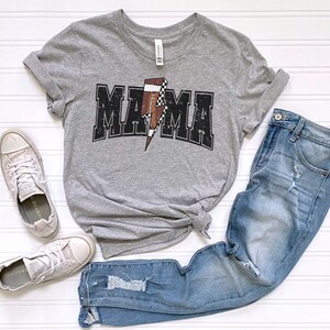 May include: Gray t-shirt with a black and white football graphic and the word "MAMA" in black letters. The shirt is paired with blue jeans and white sneakers.