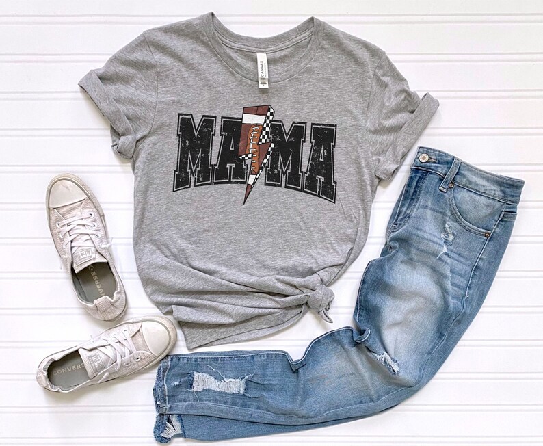 May include: Gray t-shirt with a black and white football graphic and the word "MAMA" in black letters. The shirt is paired with blue jeans and white sneakers.