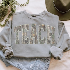 May include: A sage green sweatshirt with the word "TEACH" in large, block letters across the front. The letters are filled with a camouflage pattern. The sweatshirt has a crew neck and long sleeves.