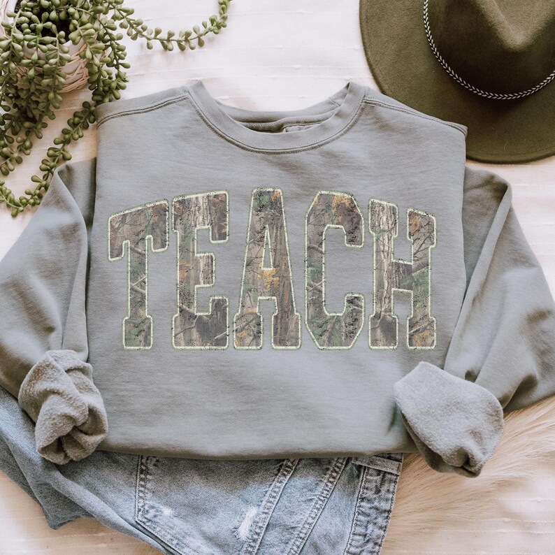 May include: A sage green sweatshirt with the word "TEACH" in large, block letters across the front. The letters are filled with a camouflage pattern. The sweatshirt has a crew neck and long sleeves.