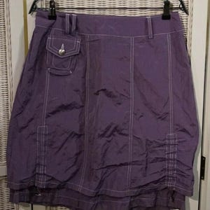 May include: A purple skirt with white stitching details and a small pocket with a heart-shaped accent. The skirt is hanging on a black hanger. The fabric appears to be a lightweight material.