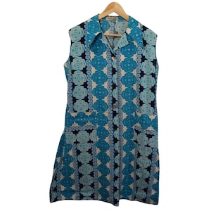 May include: A sleeveless vintage dress with a blue and white geometric pattern. The dress has a collared neckline, button-down front, and two front pockets. The dress is hanging on a wooden hanger.