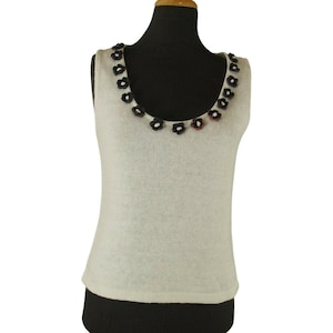 May include: A sleeveless cream-colored knit tank top featuring a scoop neckline adorned with a series of small, dark-colored floral embellishments. The garment has a simple, straight-cut silhouette and a fine, lightweight knit texture throughout.