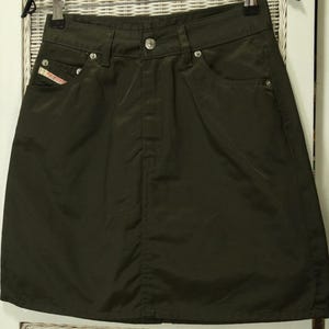 May include: Dark olive green skirt with a classic five-pocket design. Features a button closure and metal rivets. The skirt has a slight A-line shape and is made of a smooth fabric. A small red and white logo is visible on the front pocket.