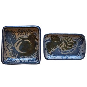 May include: Two ceramic dishes with a speckled beige base and dark blue glaze. The square dish features a bird illustration, while the rectangular dish displays a bird in flight. Both pieces have dark blue edges and a hand-painted aesthetic.