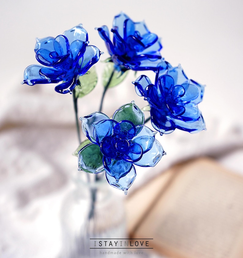 May include: A bouquet of blue glass flowers in a clear glass vase. The flowers have multiple petals and green leaves. The text "I STAY IN LOVE handmade with love" is at the bottom of the image.