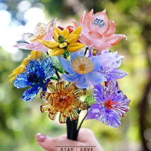 May include: A vibrant bouquet of glass flowers in pink, yellow, blue, and purple. The flowers have detailed designs and are held together with a dark stem. The text "ISTAYINLOVE handmade with love" is at the bottom.