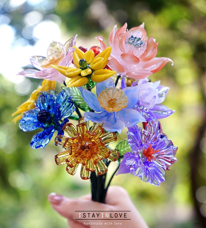 May include: A vibrant bouquet of glass flowers in pink, yellow, blue, and purple. The flowers have detailed designs and are held together with a dark stem. The text "ISTAYINLOVE handmade with love" is at the bottom.