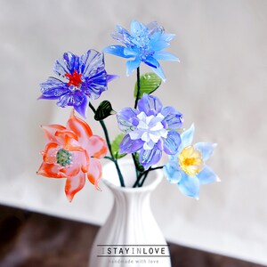 May include: A white vase displays a bouquet of glass flowers in shades of blue, purple, and orange, with green stems and leaves. The text "I STAY IN LOVE" is at the base of the vase.