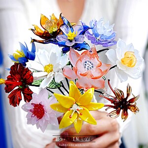 May include: A vibrant bouquet of handmade glass flowers in a variety of colours, including red, yellow, orange, blue, and white. The flowers are arranged together, creating a colourful display. The text "I STAY IN LOVE" is visible at the bottom.