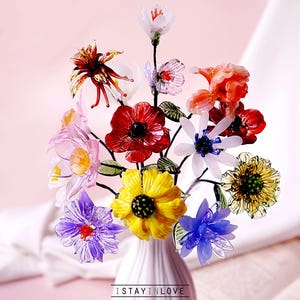 May include: A vibrant bouquet of glass flowers in a white vase. The arrangement features red, yellow, purple, and blue blooms with green leaves. The vase is on a pink surface. Text at the bottom reads "ISTAYINLOVE handmade with love."