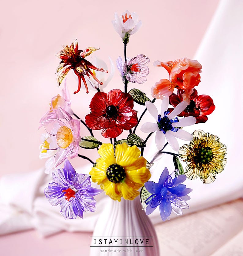 May include: A vibrant bouquet of glass flowers in a white vase. The arrangement features red, yellow, purple, and blue blooms with green leaves. The vase is on a pink surface. Text at the bottom reads "ISTAYINLOVE handmade with love."