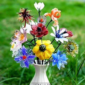 May include: A vibrant bouquet of glass flowers in a white vase. The flowers are in various colours, including red, yellow, and blue, with detailed designs. The vase is set against a blurred green backdrop. Text at the bottom reads "ISTAYINLOVE handmade with love."