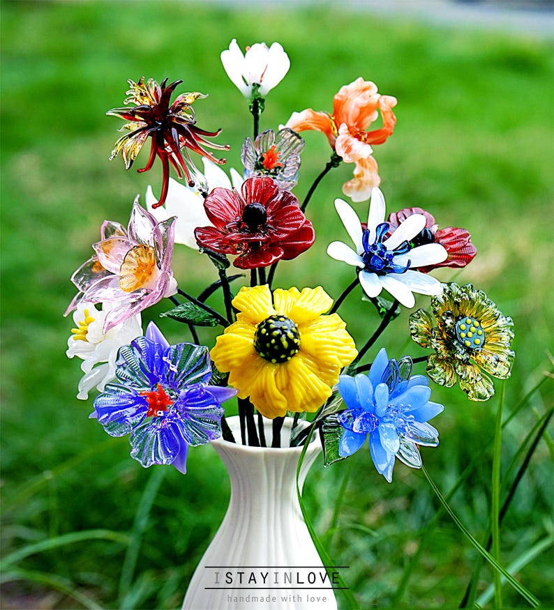 May include: A vibrant bouquet of glass flowers in a white vase. The flowers are in various colours, including red, yellow, and blue, with detailed designs. The vase is set against a blurred green backdrop. Text at the bottom reads "ISTAYINLOVE handmade with love."