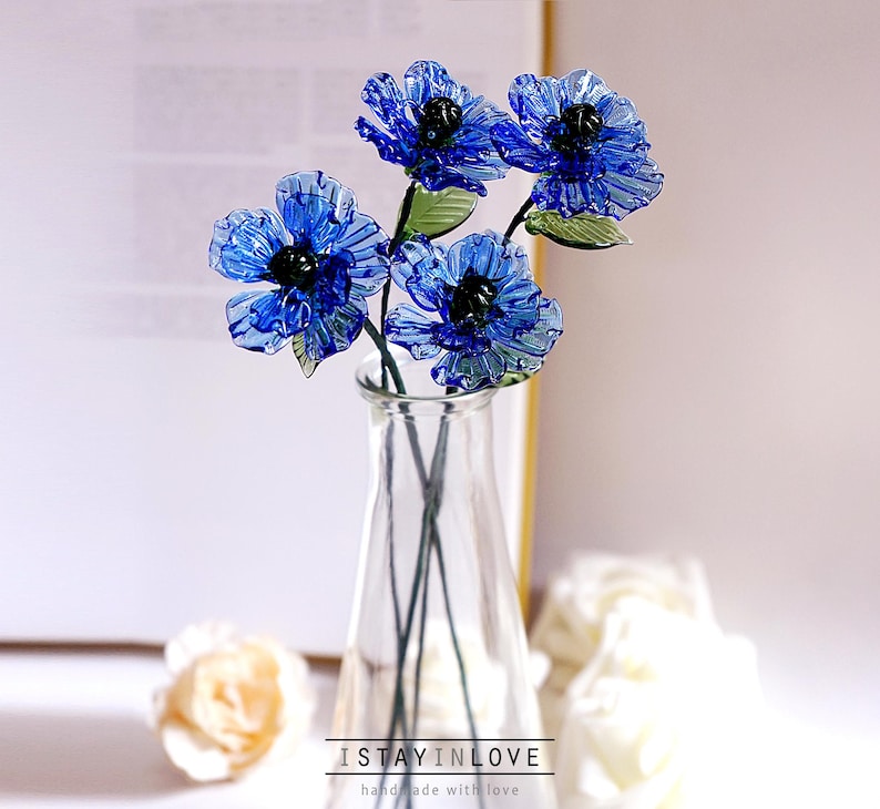 May include: A clear glass vase holding a bouquet of blue glass flowers with black centres and green leaves. The flowers are in full bloom. The text "I STAY IN LOVE" is at the bottom of the image.
